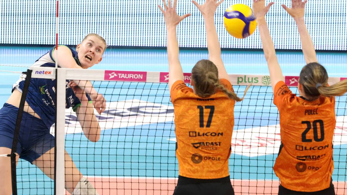 Siatkarka w niebieskim stroju blokuje piłkę na tle siatki, po drugiej stronie siatki dwie siatkarki w pomarańczowych strojach pr&oacute;bują odbić piłkę.