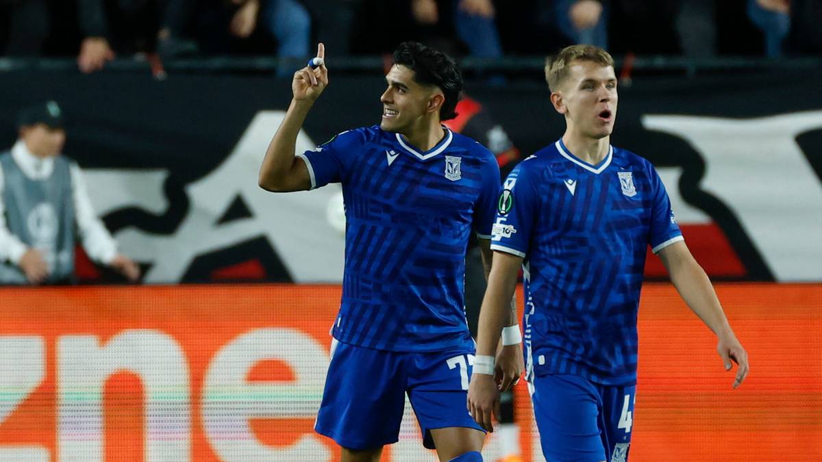 Dw&oacute;ch piłkarzy Lecha Poznań w niebieskich strojach sportowych idzie po boisku piłkarskim.