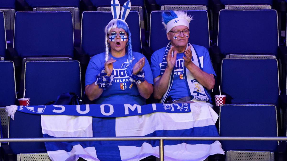 Dwoje kibiców w niebieskich koszulkach z białymi elementami, w niebiesko-białych nakryciach głowy, siedzi na trybunach, malując twarze w barwy narodowe. Przed nimi rozłożona jest flaga Finlandii.