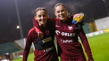 Dwie uśmiechnięte piłkarki w bordowych strojach reprezentacji Polski pozują do zdjęcia na boisku.