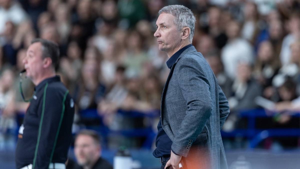 Mężczyzna w szarym garniturze z rękami na biodrach, na tle publiczności na meczu siatk&oacute;wki.