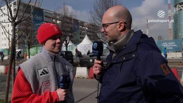 Dw&oacute;ch mężczyzn rozmawia na zewnątrz. Jeden z nich, ubrany w czerwoną bluzę i szarą kamizelkę, trzyma mikrofon. Drugi, w ciemnej kurtce i okularach, r&oacute;wnież trzyma mikrofon.