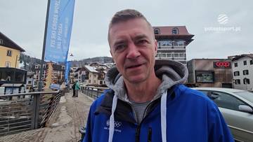 Mężczyzna w niebieskiej bluzie z kapturem, stojący na zewnątrz, z budynkami i drzewami w tle. Na jego bluzie widoczne są olimpijskie k&oacute;łka.