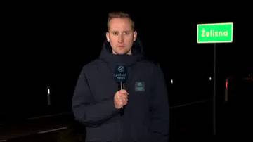 Dziennikarz z mikrofonem podczas nocnej relacji na tle drogi i tablicy z napisem "Żelazna".