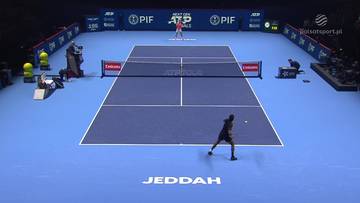 Dw&oacute;ch tenisist&oacute;w grających mecz na niebieskim korcie tenisowym. Jeden z nich, w ciemniejszym stroju, odbija piłkę w kierunku przeciwnika, kt&oacute;ry stoi bliżej siatki.