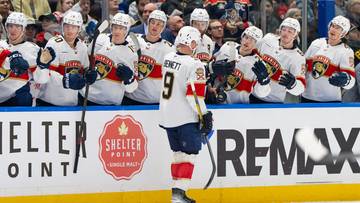 Drużyna hokejowa w białych strojach z czerwonymi i złotymi akcentami stoi przy bandzie, z jednym graczem numer 9 odwr&oacute;conym tyłem do widza na pierwszym planie.