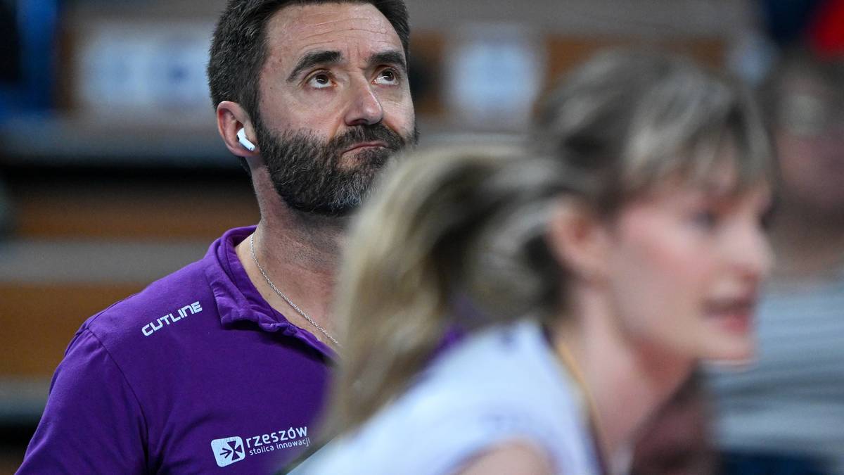 Trener siatkówki w fioletowej koszulce z białym logo, patrzący w górę, z mikrofonem w uchu.