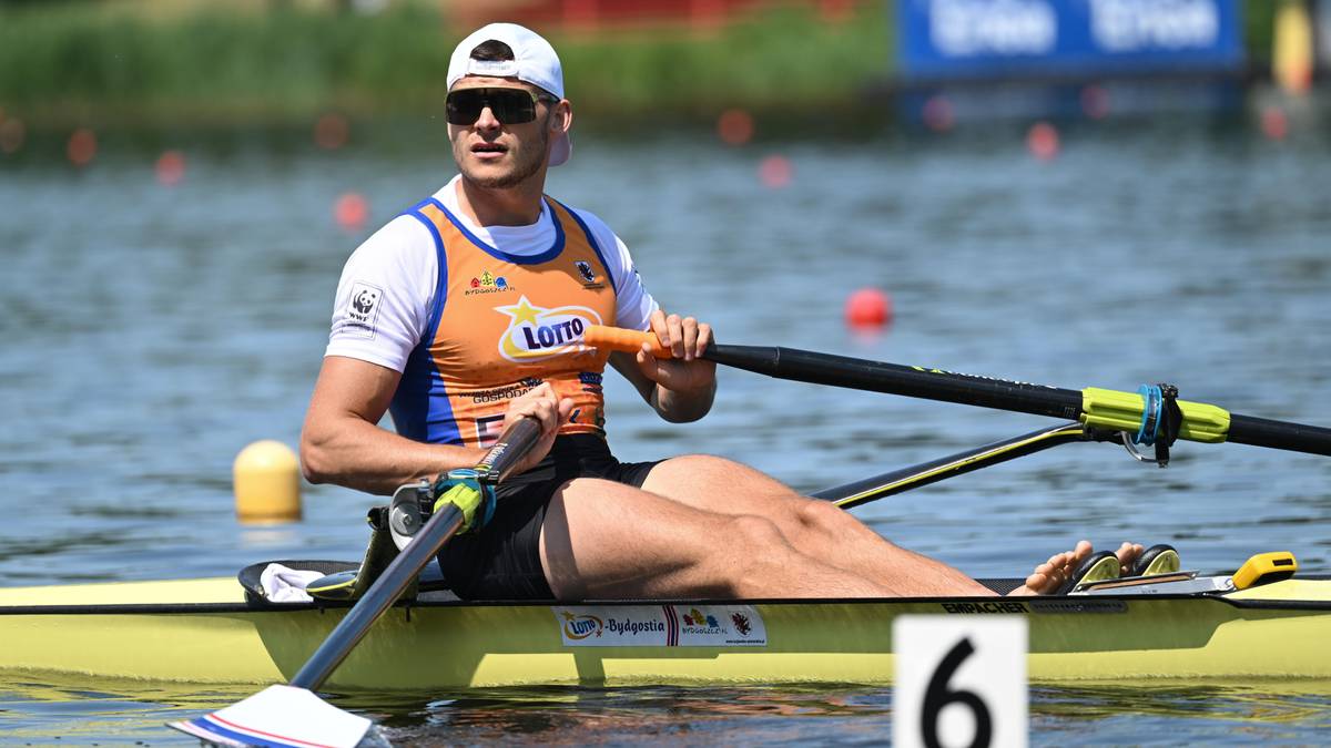 Mężczyzna wioślarz w czapce i okularach przeciwsłonecznych, ubrany w białą koszulkę z logo LOTTO i pomarańczową kamizelkę, wiosłuje w łodzi podczas zawod&oacute;w.