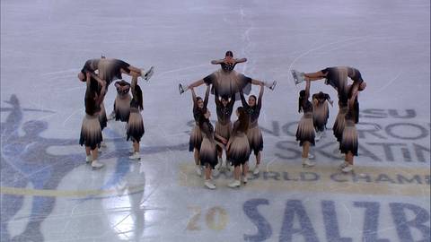 Grupa łyżwiarzy synchronicznych w brązowo-beżowych strojach wykonuje figury na lodowisku.