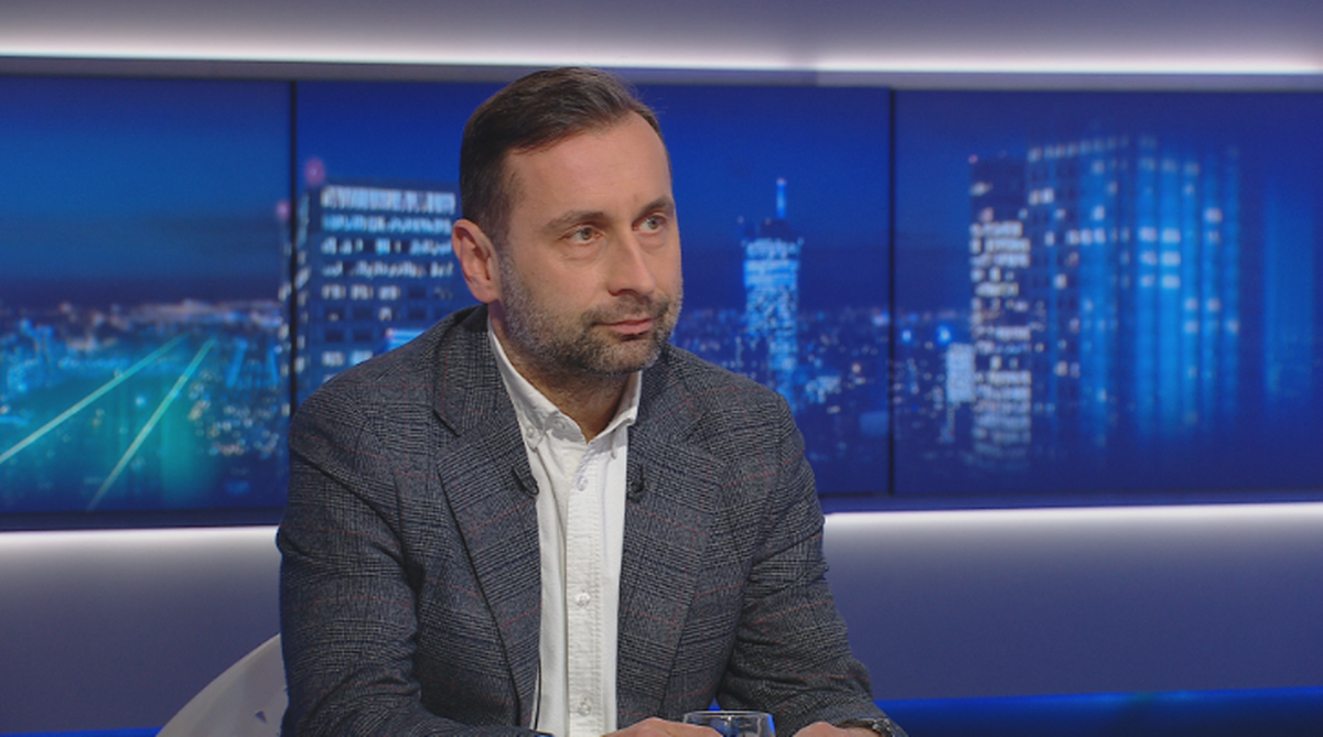 Adam Balcer w studiu telewizyjnym, siedzi przed panelem z ekranami wyświetlającymi nocną panoramę miasta.