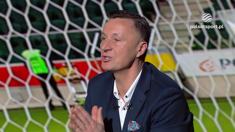 Tomasz Hajto w eleganckim garniturze, siedzący przed bramką piłkarską i gestykulujący rękami.