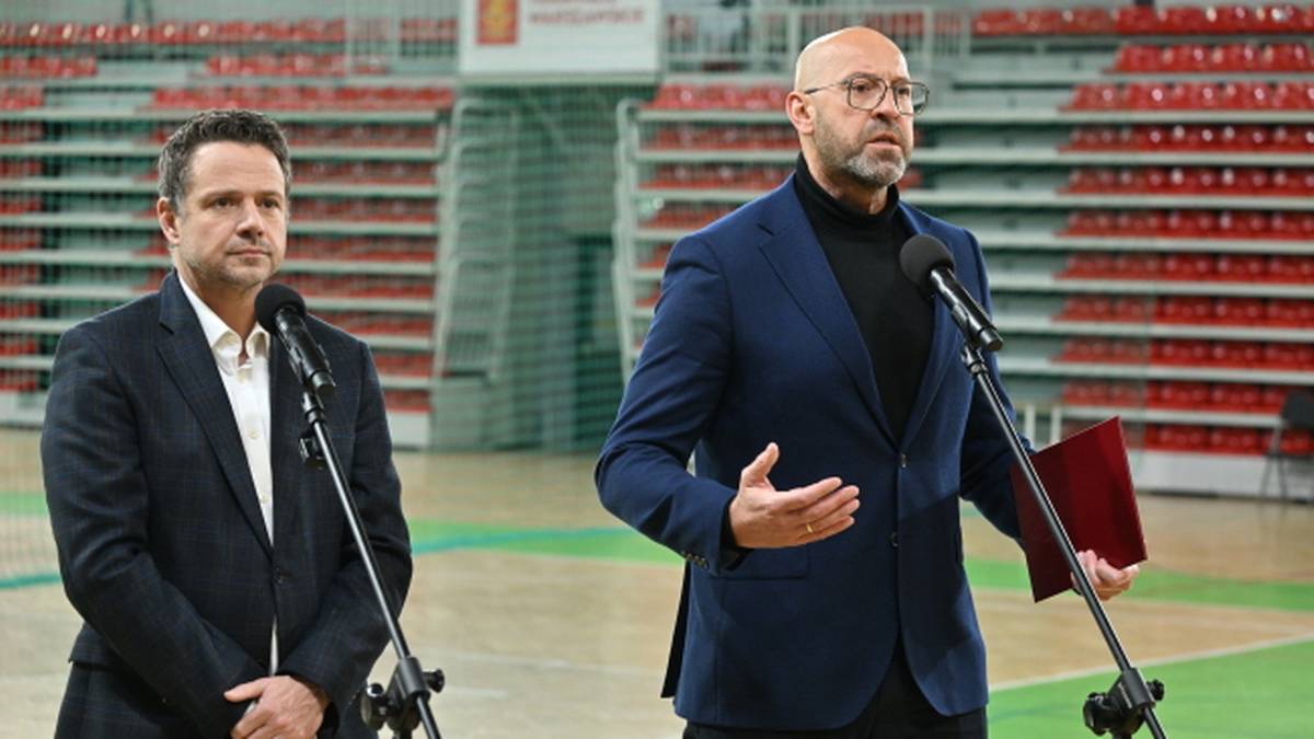 Dwóch mężczyzn w garniturach przemawia do mikrofonów w hali sportowej.