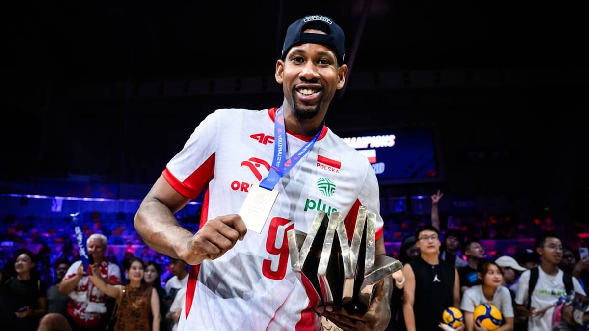 Siatkarz reprezentacji Polski w białej koszulce z czerwonymi elementami, trzymający medal i trofeum, uśmiecha się do obiektywu.