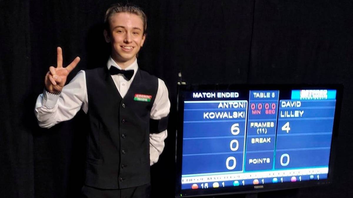 Młody mężczyzna w kamizelce i muszce pokazuje dwa palce do kamery.