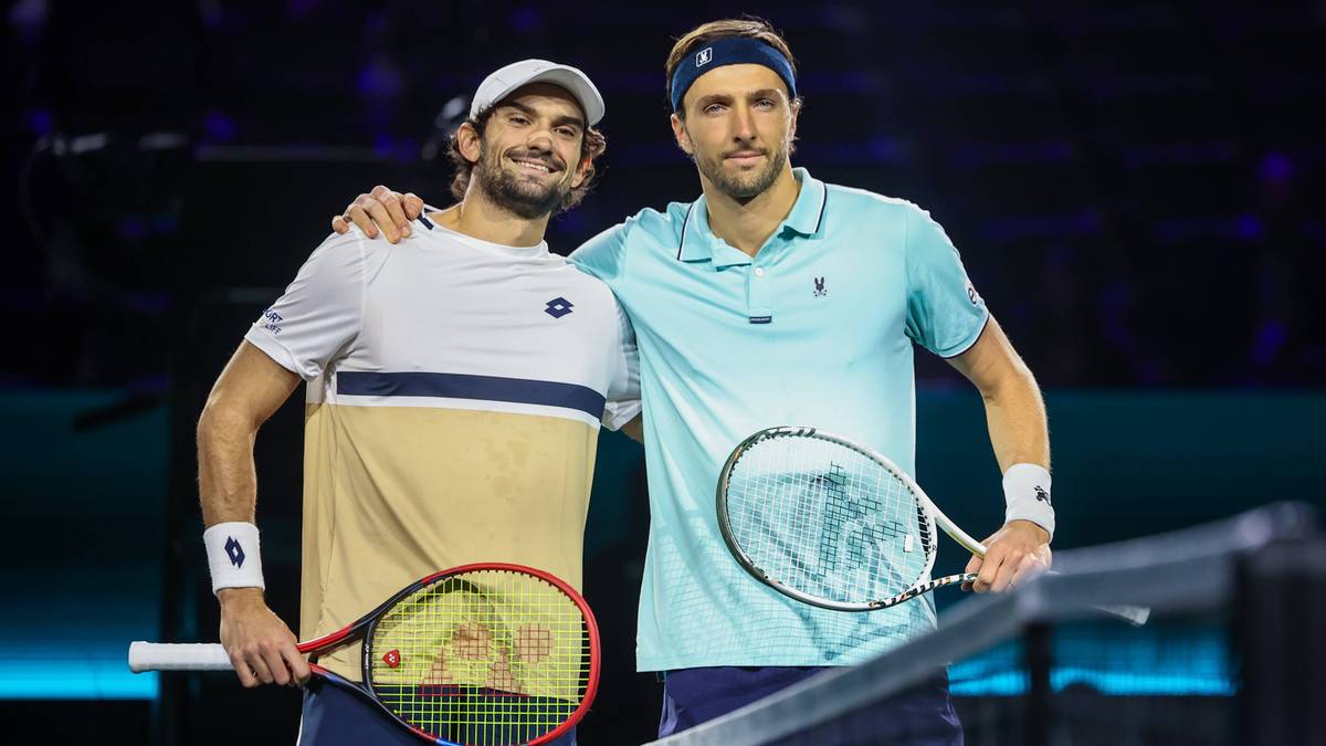 Dw&oacute;ch mężczyzn w strojach tenisowych stoi przy siatce tenisowej, jeden trzyma rakietę.