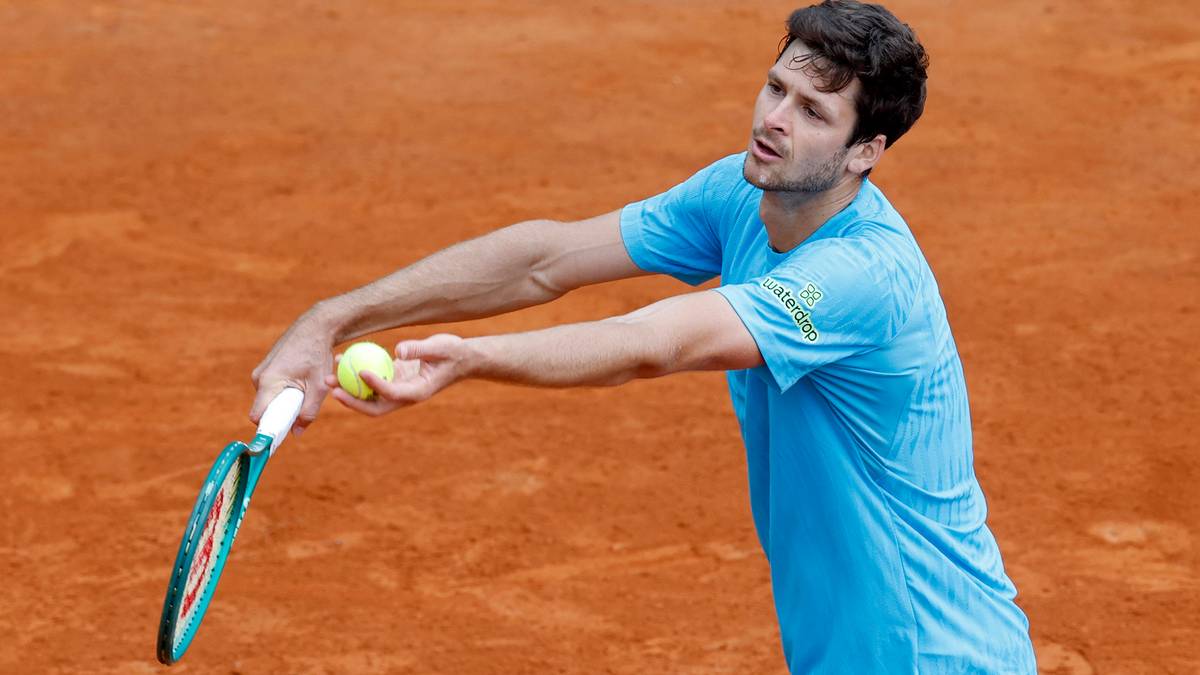 Tenisista w niebieskiej koszulce serwuje piłkę na korcie ziemnym.