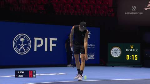 Tenisista Alexander Blockx podczas meczu, pochylony, aby podnieść piłkę tenisową. W tle widać tablicę wynik&oacute;w.