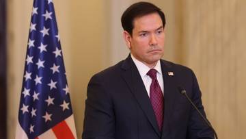 Marco Rubio w garniturze i krawacie stojący na tle flagi USA.