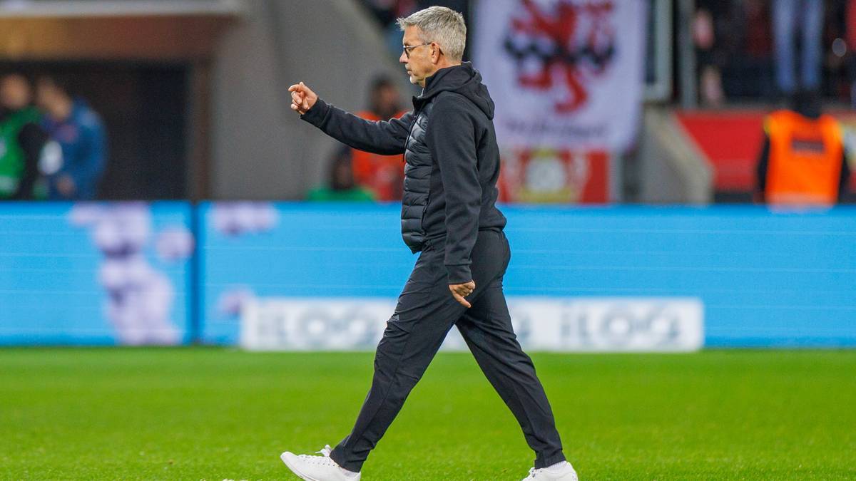 Trener piłkarski w czarnym dresie stoi na boisku, wskazując palcem w lewo.