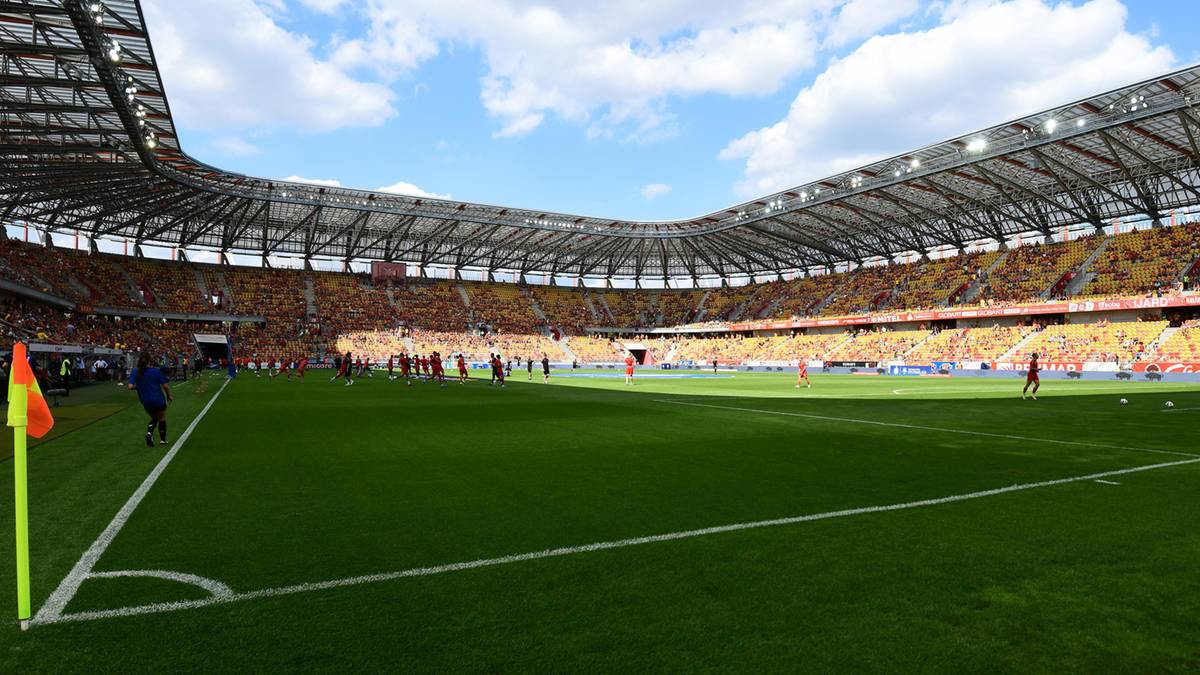 Widok na zielone boisko piłkarskie stadionu z trybunami wypełnionymi kibicami.