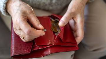 Dłonie starszej osoby trzymające czerwony portfel z monetami, jedna moneta wyjmowana jest z portfela.