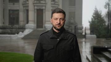 Wołodymyr Zełenski, prezydent Ukrainy, stoi przed budynkiem w deszczowy dzień.