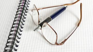 Okulary i długopis leżą na notatniku z kratkowanymi kartkami.