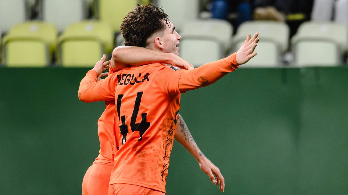 Dwóch piłkarzy w pomarańczowych strojach obejmuje się na boisku, świętując zwycięstwo. Widoczne numery na koszulkach.