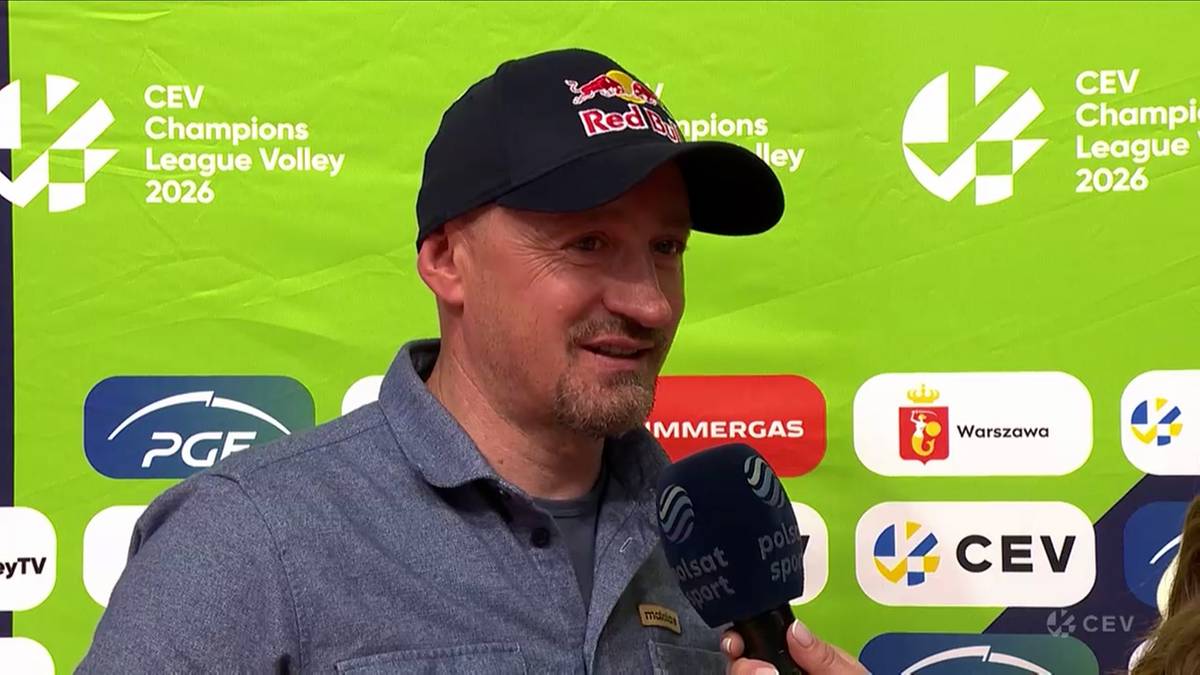 Adam Małysz, ubrany w szarą koszulę i czarną czapkę z logo Red Bull, uśmiecha się podczas wywiadu przed banerem Ligi Mistrz&oacute;w CEV.