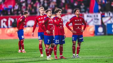 Pięciu piłkarzy w czerwonych strojach z niebieskimi detalami stoi na zielonej murawie stadionu piłkarskiego, patrząc w r&oacute;żnych kierunkach.