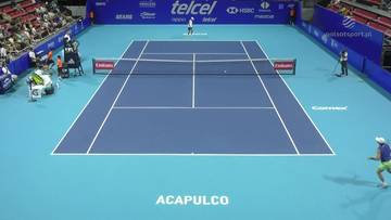 Kort tenisowy podczas meczu, widoczni dwaj zawodnicy w białych strojach i trybuny z widzami.