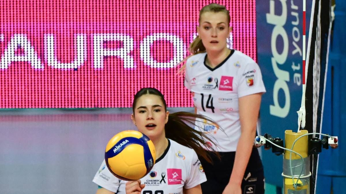Dwie siatkarki w białych strojach sportowych. Jedna z nich trzyma piłkę do siatkówki, druga stoi za nią z numerem 14.