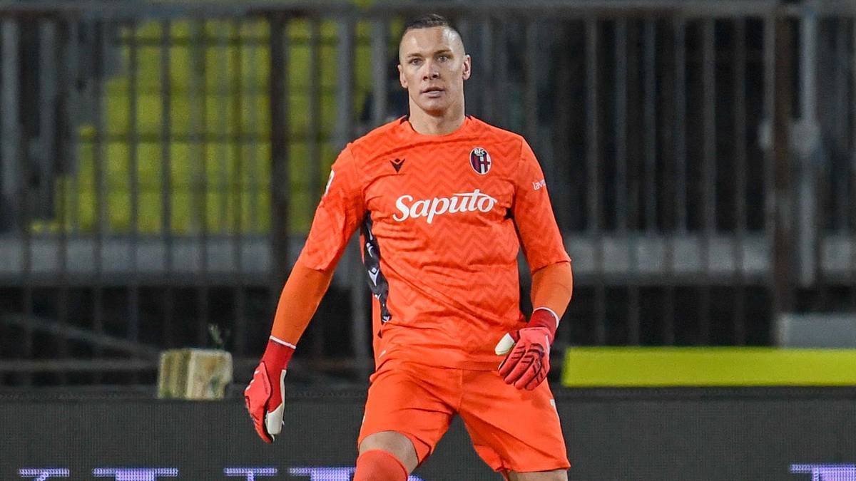 Bramkarz w pomarańczowym stroju z rękawicami bramkarskimi na boisku piłkarskim.