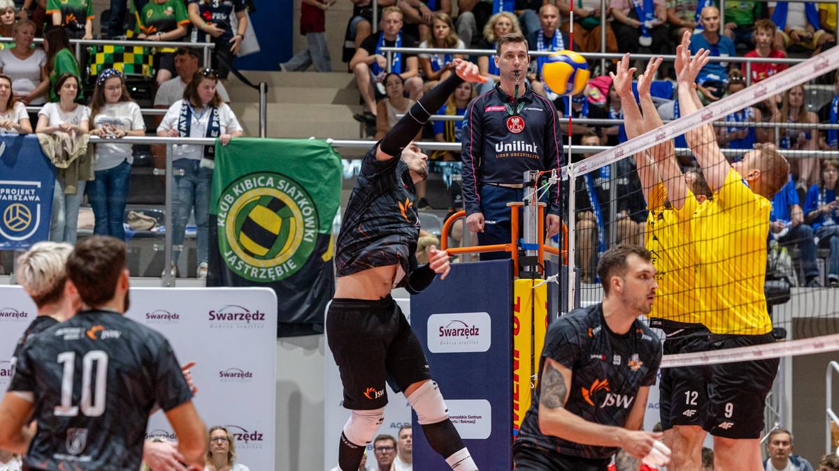 Siatkarz w czarnej koszulce atakuje piłkę nad siatką, blokowany przez dwóch zawodników w żółtych koszulkach.