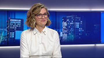 Kobieta z okularami i kr&oacute;tkimi blond włosami, ubrana w białą koszulę, uśmiecha się.