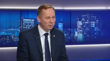 Mężczyzna w ciemnym garniturze i białej koszuli z krawatem, siedzi i m&oacute;wi. W tle za nim znajduje się ekran z widokiem nocnego miasta.