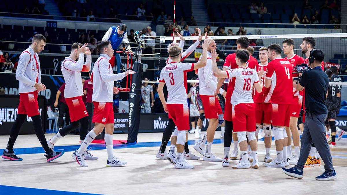 Grupa siatkarzy w białych koszulkach i czerwonych spodenkach na boisku.