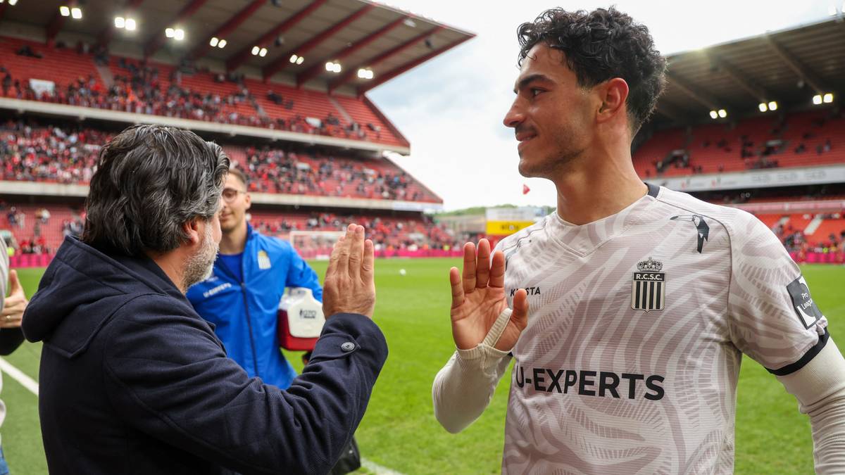 Piłkarz w białej koszulce z czarnymi akcentami i napisem "EXPERTS" po prawej stronie, rozmawia z mężczyzną po lewej, unosząc obie dłonie w geście powitania lub gratulacji. W tle widać trybuny stadionu wypełnione widzami.