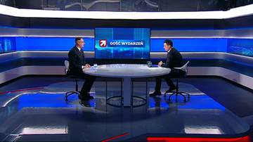 Dw&oacute;ch mężczyzn w garniturach siedzi naprzeciwko siebie przy okrągłym stole w studiu telewizyjnym. Na ekranie za nimi widnieje logo programu.