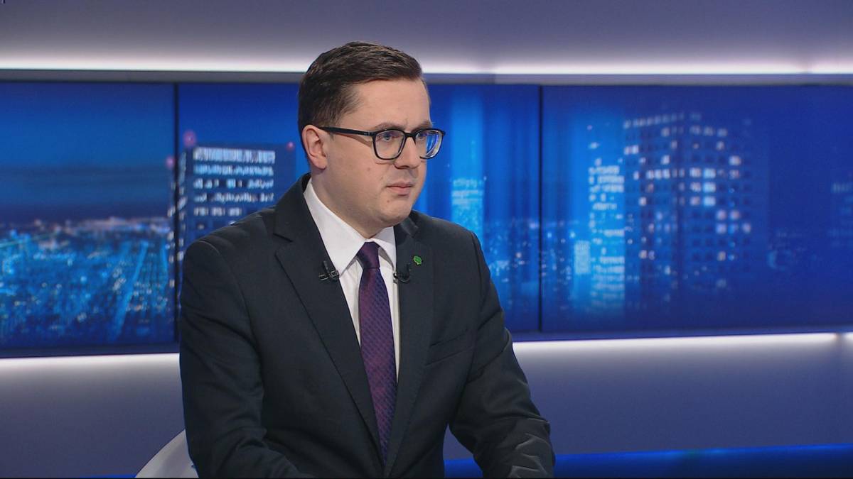 Mężczyzna w okularach i ciemnym garniturze siedzi przed ekranem z widokiem na nocne miasto.