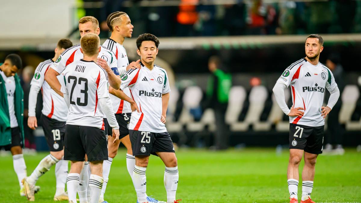 Piłkarze Legii Warszawa w białych strojach z czarnymi elementami, stojący na boisku piłkarskim.