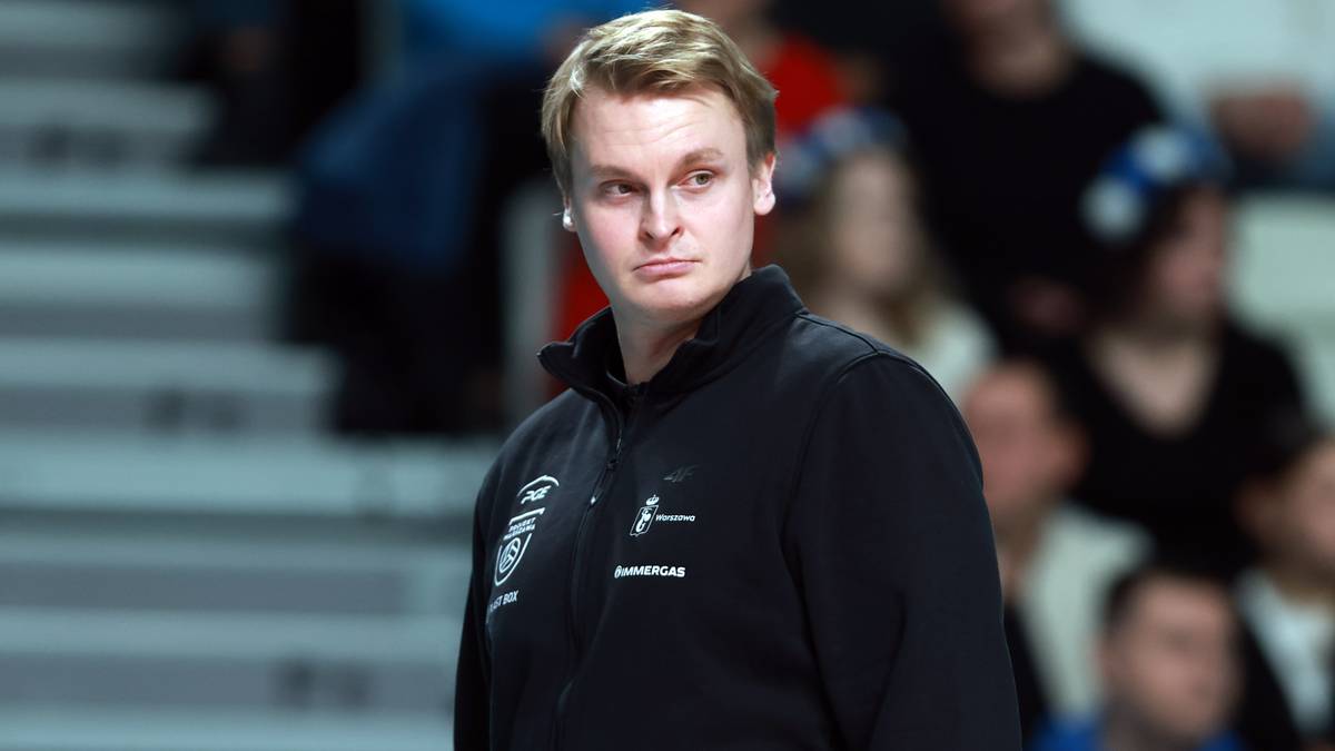 Młody mężczyzna z jasnymi włosami, ubrany w czarną bluzę z białymi logotypami, patrzy w bok z poważnym wyrazem twarzy.