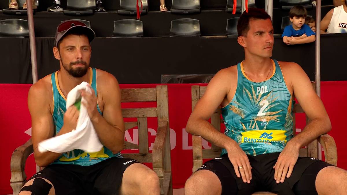 Dw&oacute;ch siatkarzy plażowych siedzi na ławce, jeden z nich wyciera twarz ręcznikiem.