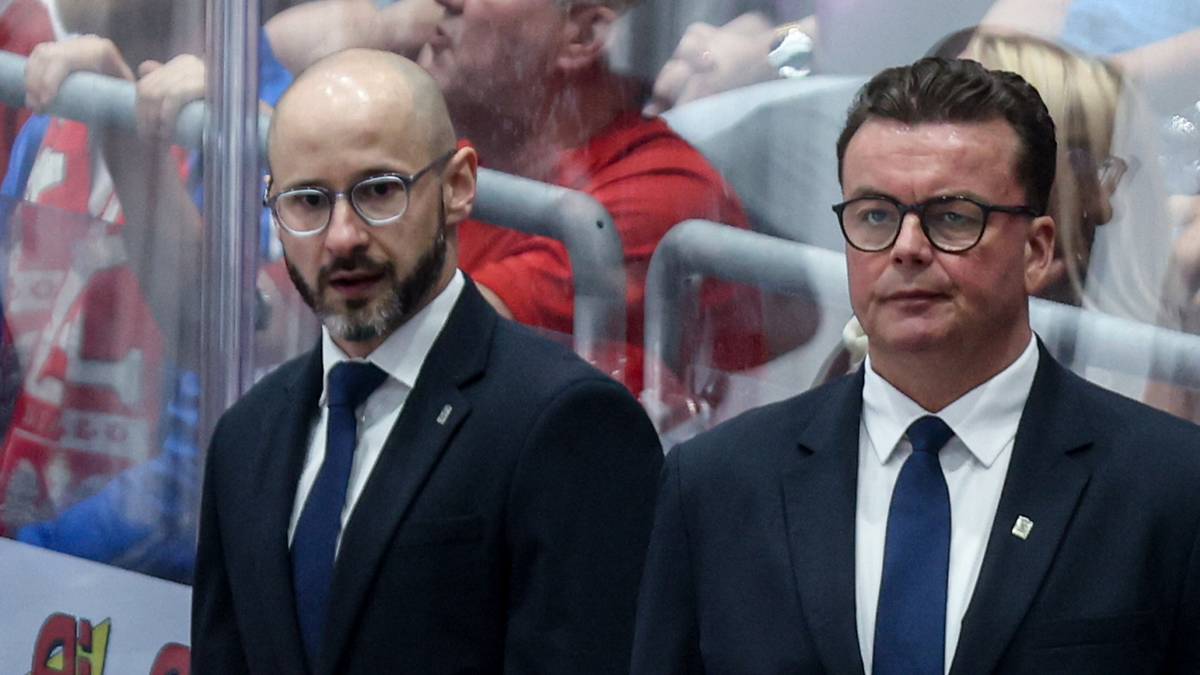 Dw&oacute;ch mężczyzn w garniturach stoi za ławką rezerwową hokejową, obserwując graczy w czerwonych kaskach.