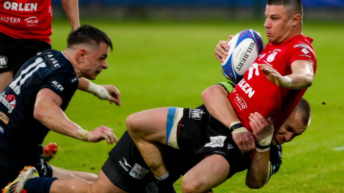 Dwóch zawodników drużyny w czarnych strojach próbuje zatrzymać zawodnika w czerwonym stroju, który biegnie z piłką do rugby.