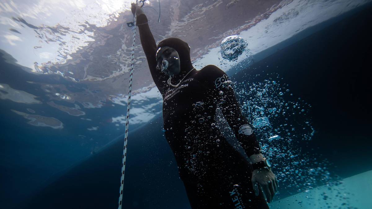 Nurkini w kombinezonie do freedivingu i masce schodzi pod wodę, trzymając się liny. Widoczne są bąbelki powietrza wokół nurka i odbicia światła od powierzchni wody.