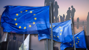 Flagi Unii Europejskiej na pierwszym planie, w tle zamazane sylwetki ludzi i budynk&oacute;w, symbolizujące członkostwo Polski w UE.