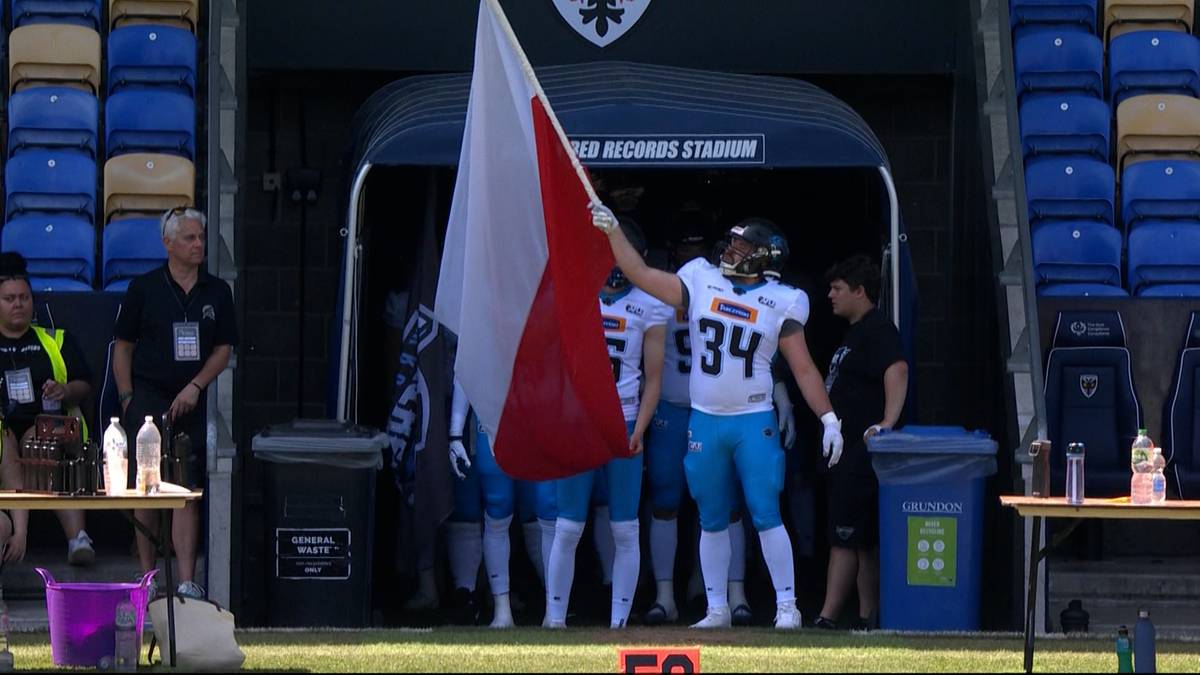 Amerykański futbolista w białej koszulce z numerem 34 i niebieskich spodenkach unosi biało-czerwoną flagę.