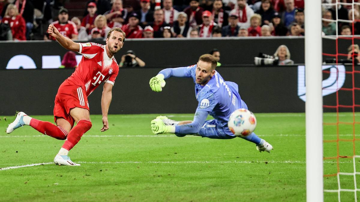 Piłkarz w czerwonej koszulce strzela gola, a bramkarz w niebieskim stroju pr&oacute;buje go obronić.