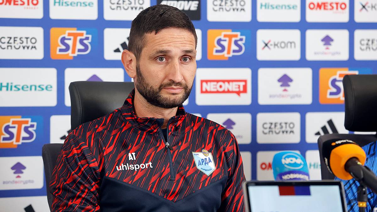 Portret mężczyzny w czarno-czerwonej koszulce polo z logo "uhlsport" i symbolem "AT", siedzącego przed ścianą z logo sponsorów.